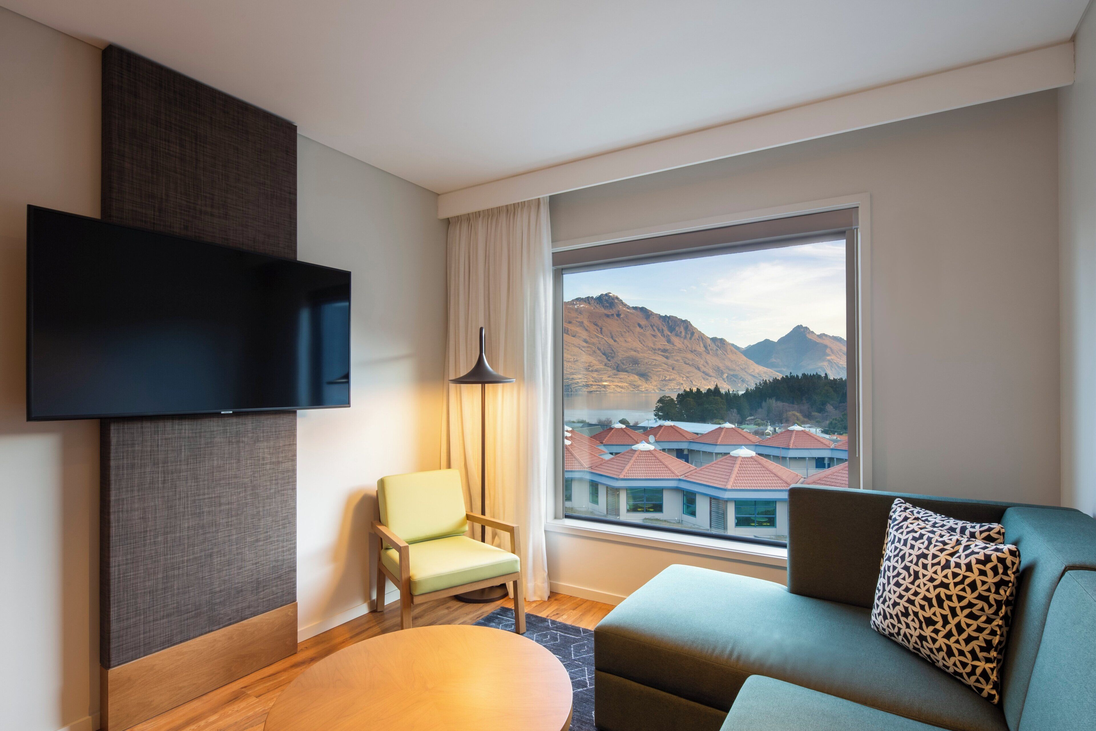 Une chambre d’hôtel moderne équipée d’une télévision à écran plat, d’un canapé, d’un fauteuil et d’une grande fenêtre donnant sur les montagnes.