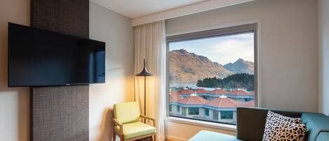 Habitación de hotel moderna con una televisión de pantalla plana, un sofá, una silla y un amplio ventanal con vistas a la montaña.