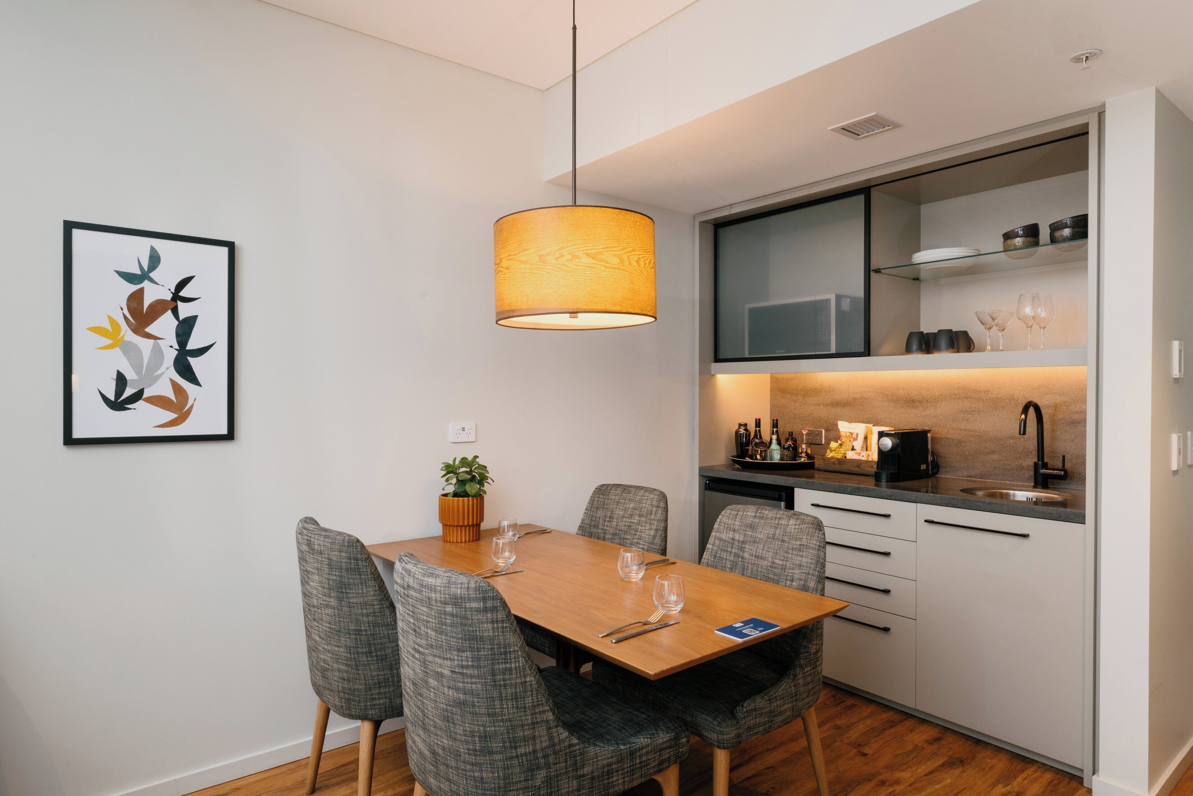 A modern kitchen with a dining area, featuring a wooden table, grey upholstered chairs, and a pendant light.