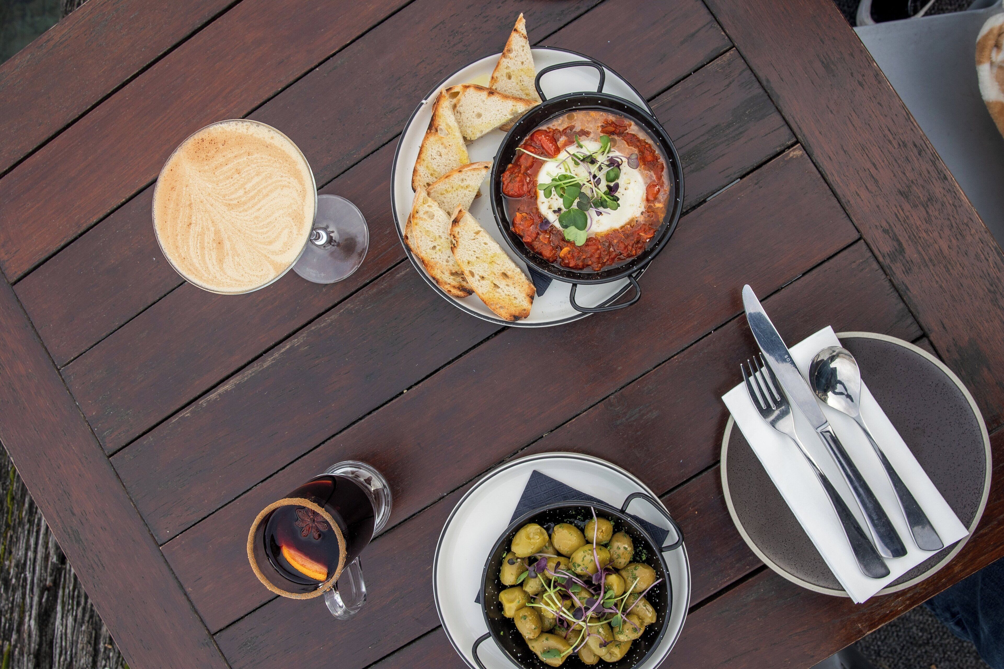 Une table en bois sur laquelle se trouvent un bol d’olives, une tasse de café et une assiette de pain.
