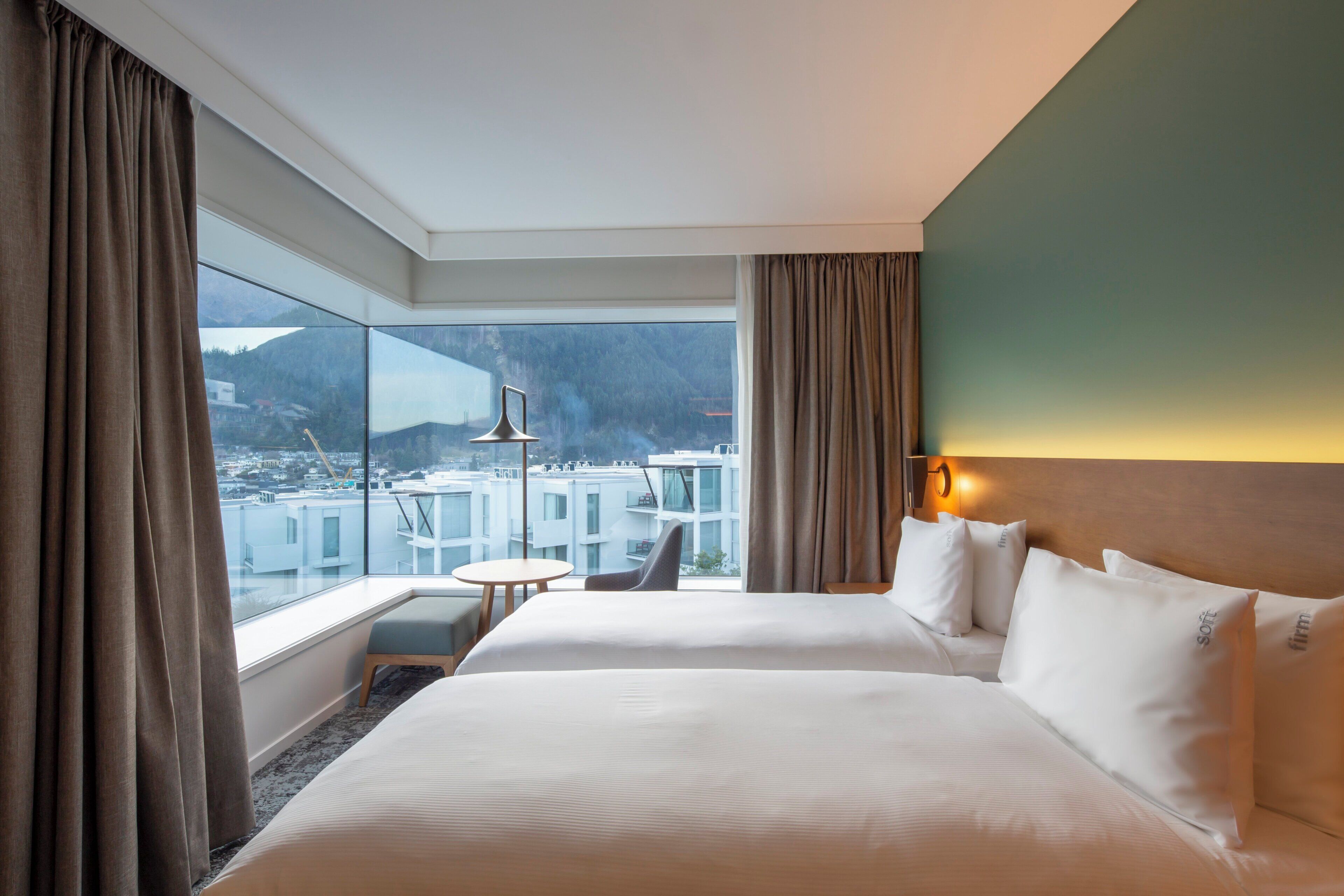 A hotel room with a large bed, a small table, a chair, and a view of a cityscape through large windows.