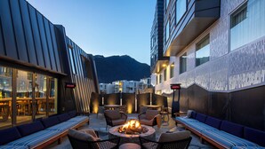 An outdoor seating area with a fire pit, lounge chairs, and tables, surrounded by modern buildings and a mountain in the background. - voco Queenstown by IHG (Queenstown)