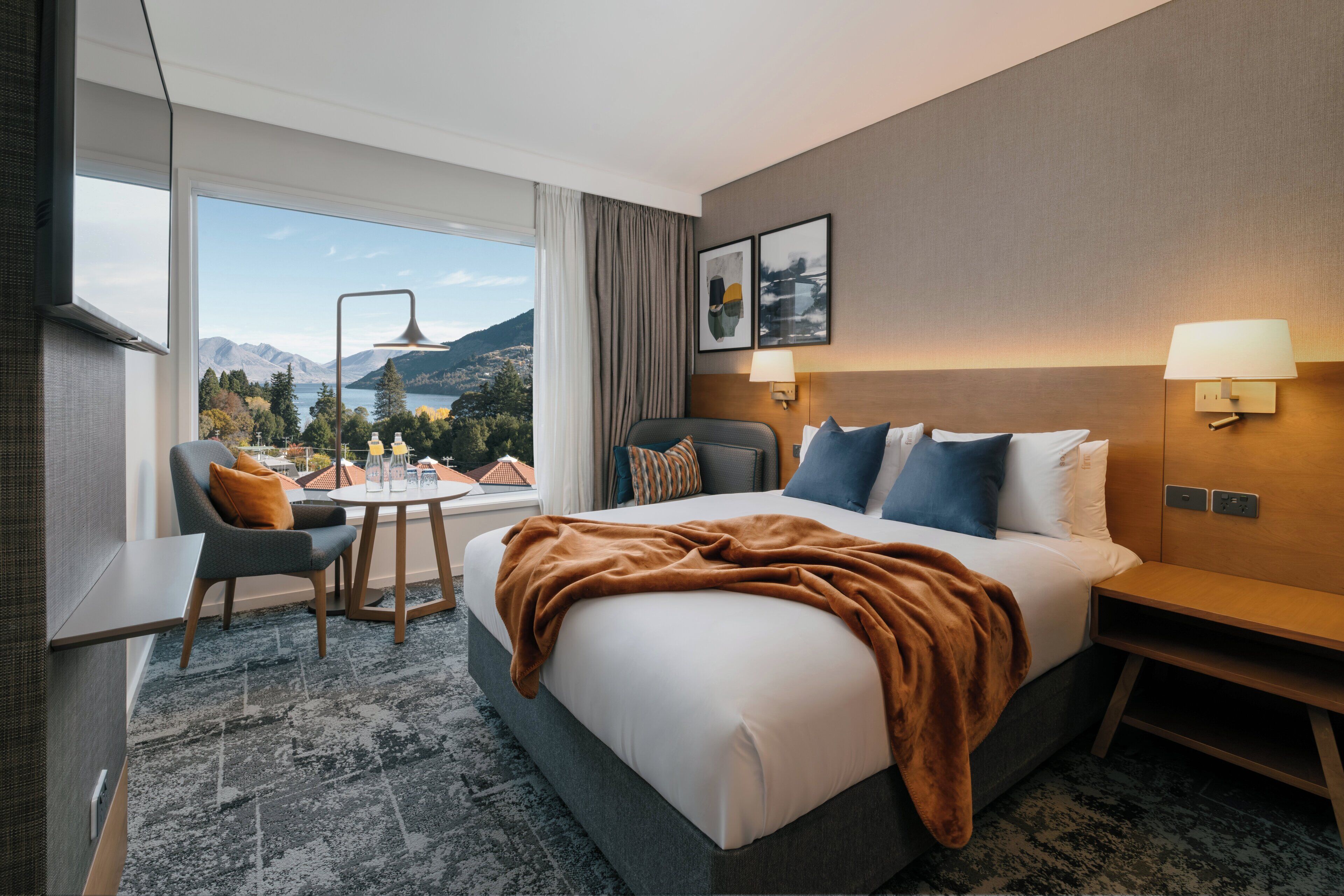 A hotel room with a bed, bedside table, lamp, chair, and a view of mountains through a large window.