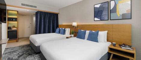 A hotel room with two beds, a desk, a small kitchenette, and artwork on the wall.