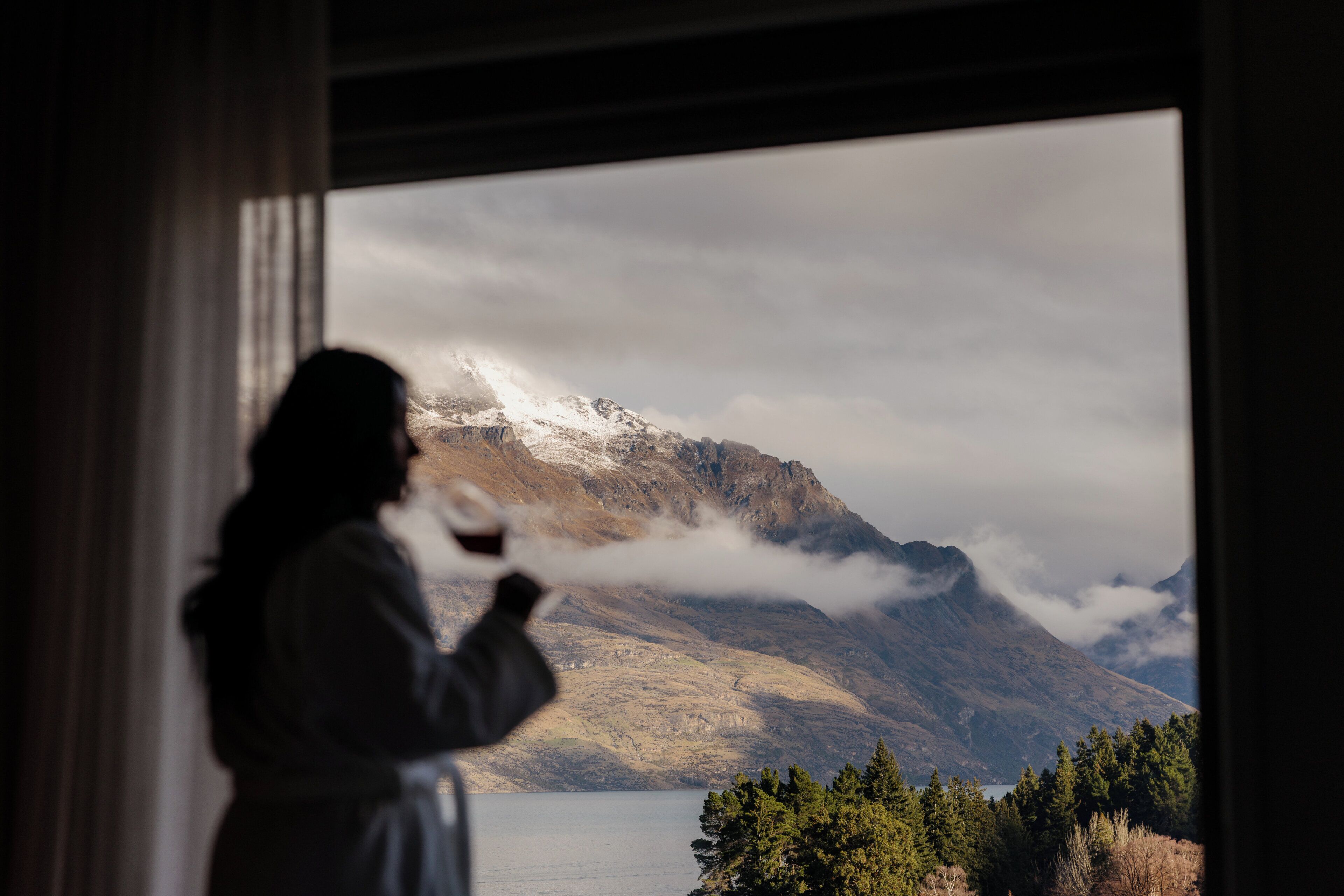 A person standing by a window with a view of mountains and a lake.