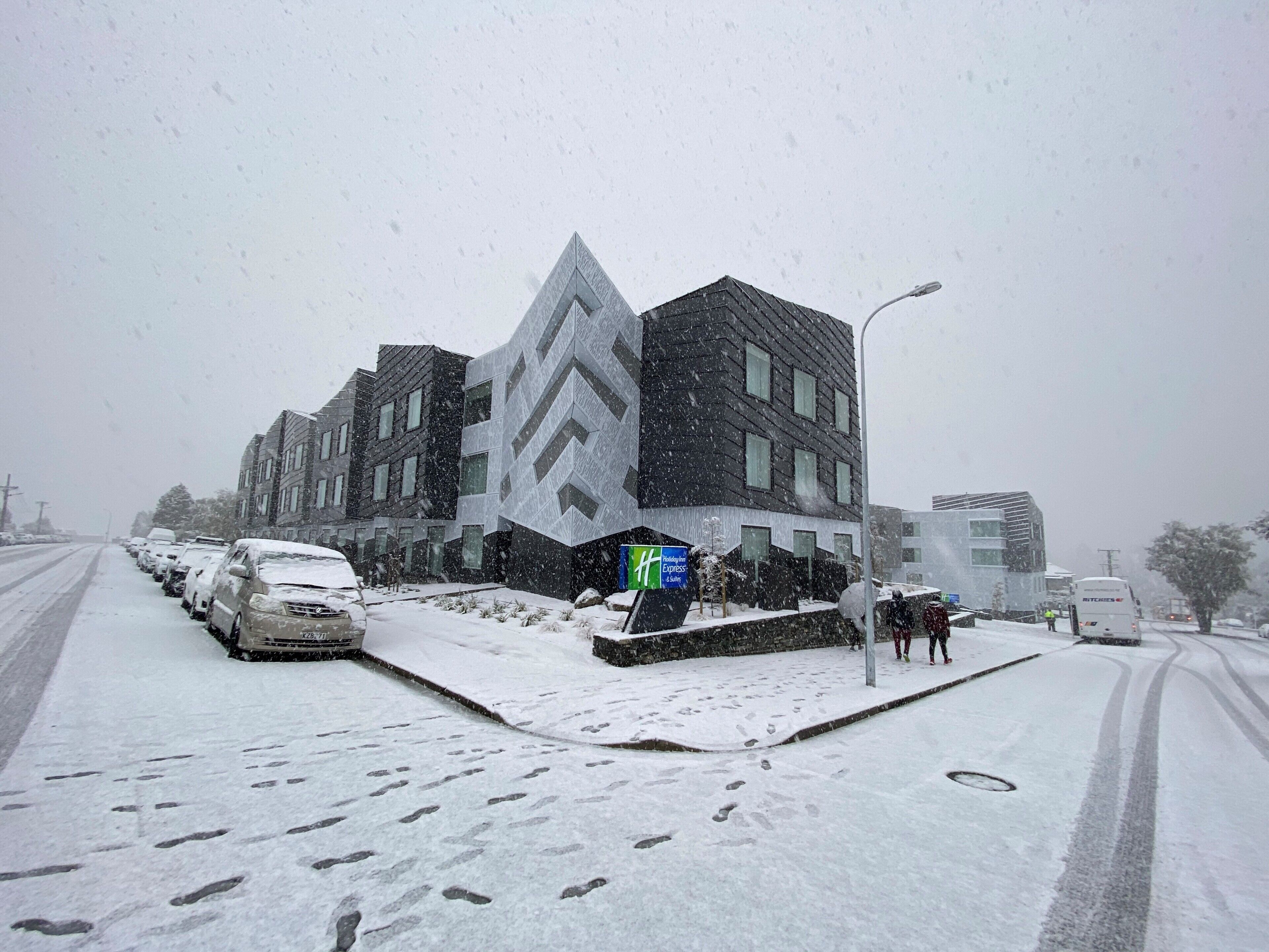 A modern building with a unique angular facade, a parking area with several cars, and a snow-covered street.