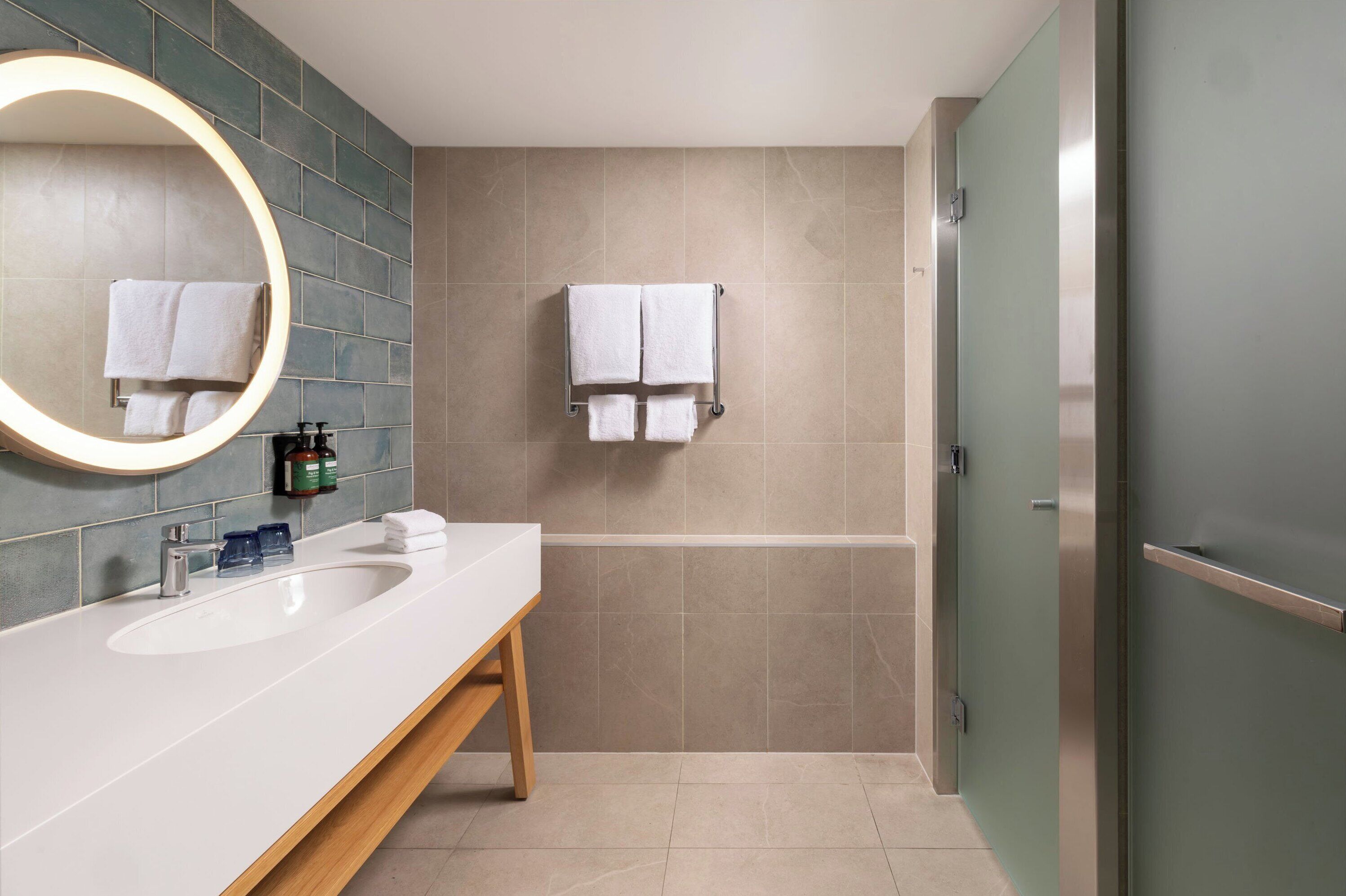 Un baño moderno con un lavamanos blanco, un espejo redondo y dos toallas colgadas en un perchero.