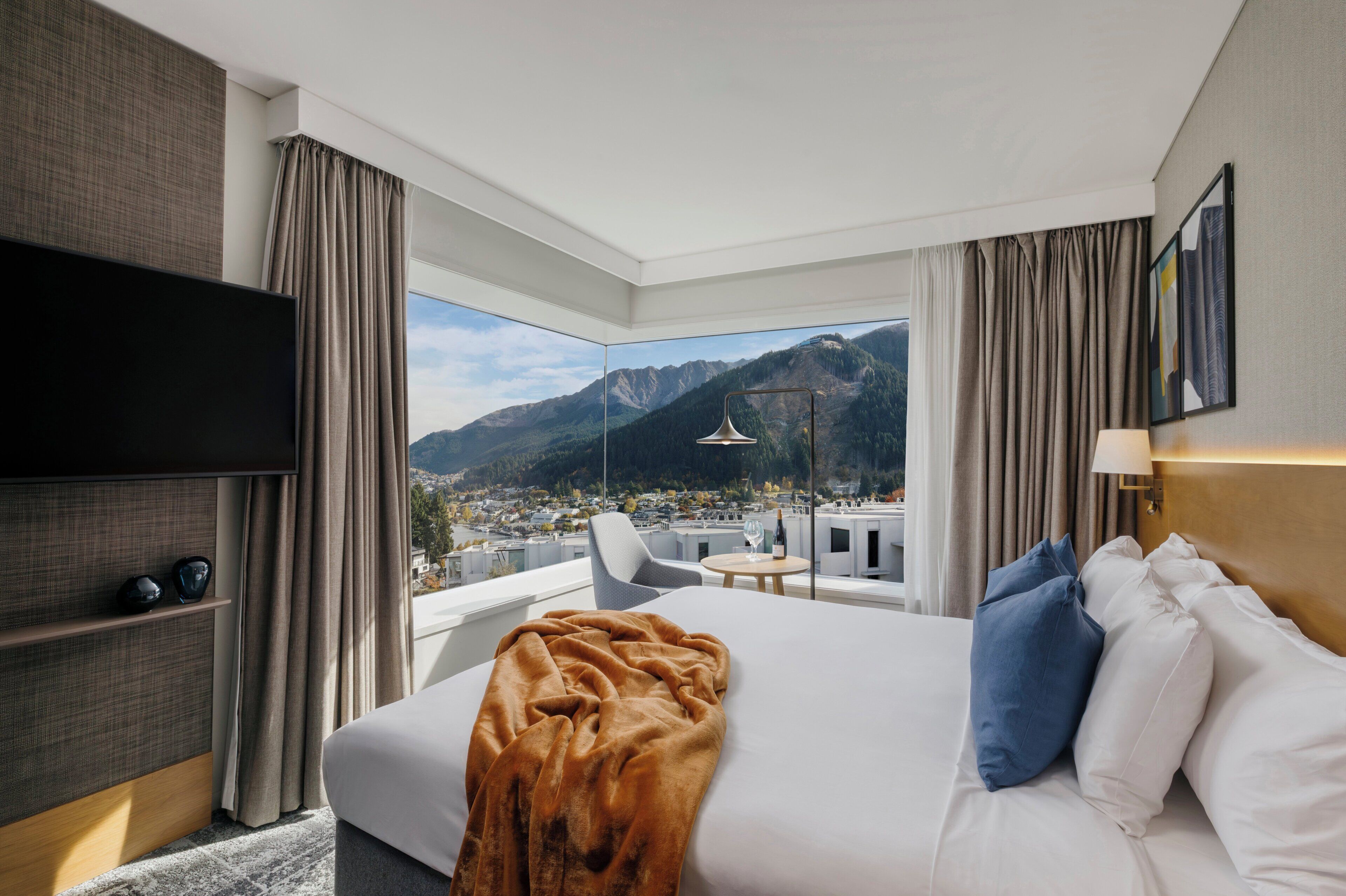 Habitación de hotel con una cama grande, televisión y vistas a las montañas y al paisaje urbano.