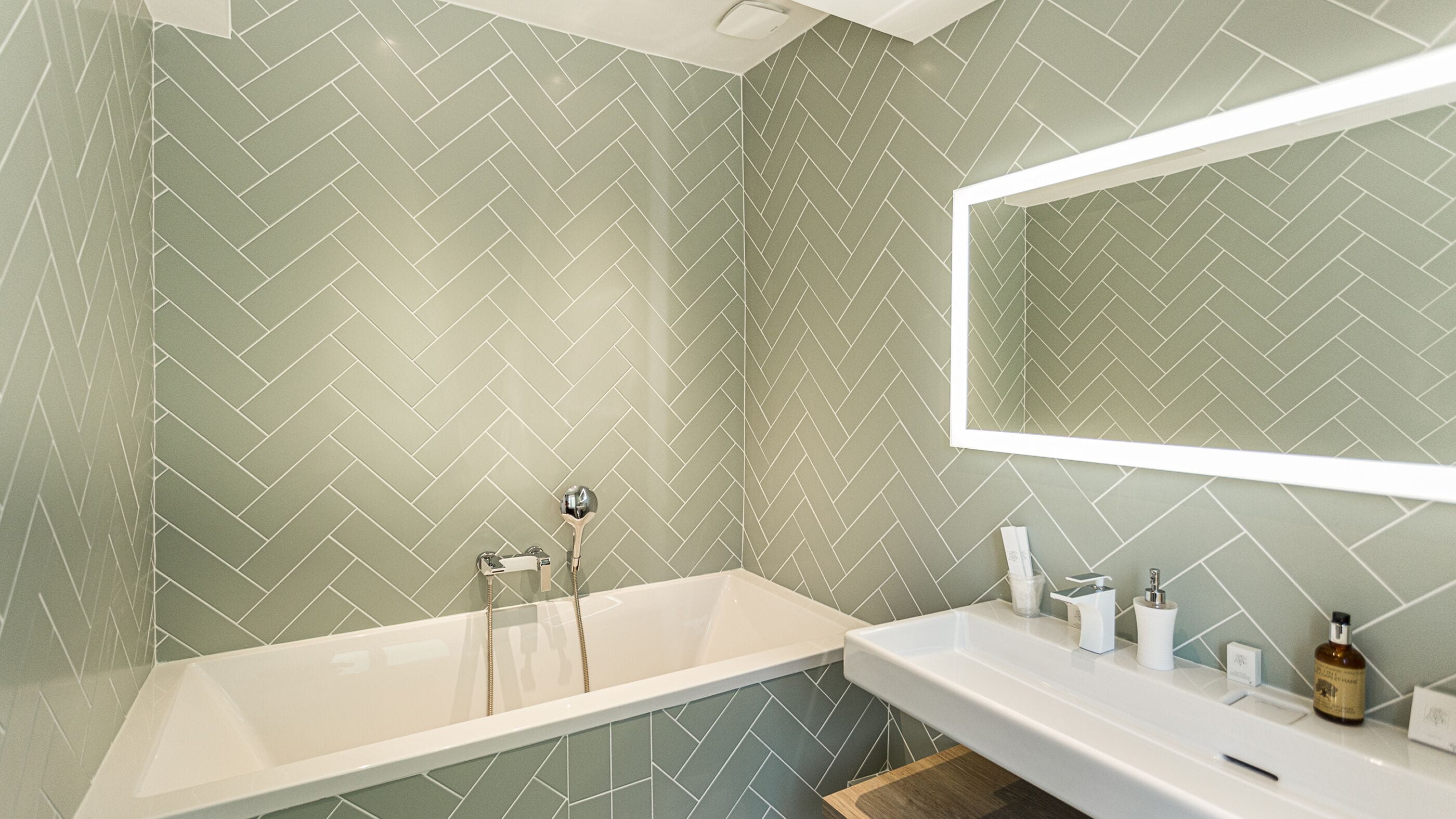 A bathroom with a herringbone-patterned tile wall, a white bathtub, a white sink, and a mirror above the sink.