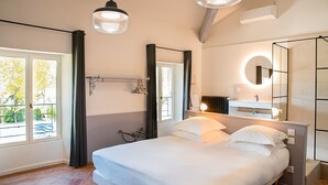 A bedroom with a bed, a window with curtains, a bedside table, a mirror, and a bathroom with a sink and mirror. - Manoir le Roure (Chateauneuf-du-Rhone)