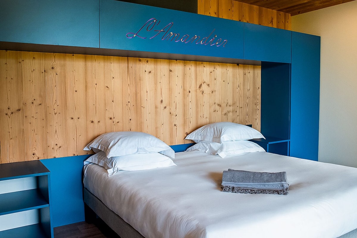 A bed with white bedding, a wooden headboard, and a blue accent wall with the text 'L'Hotelier'.