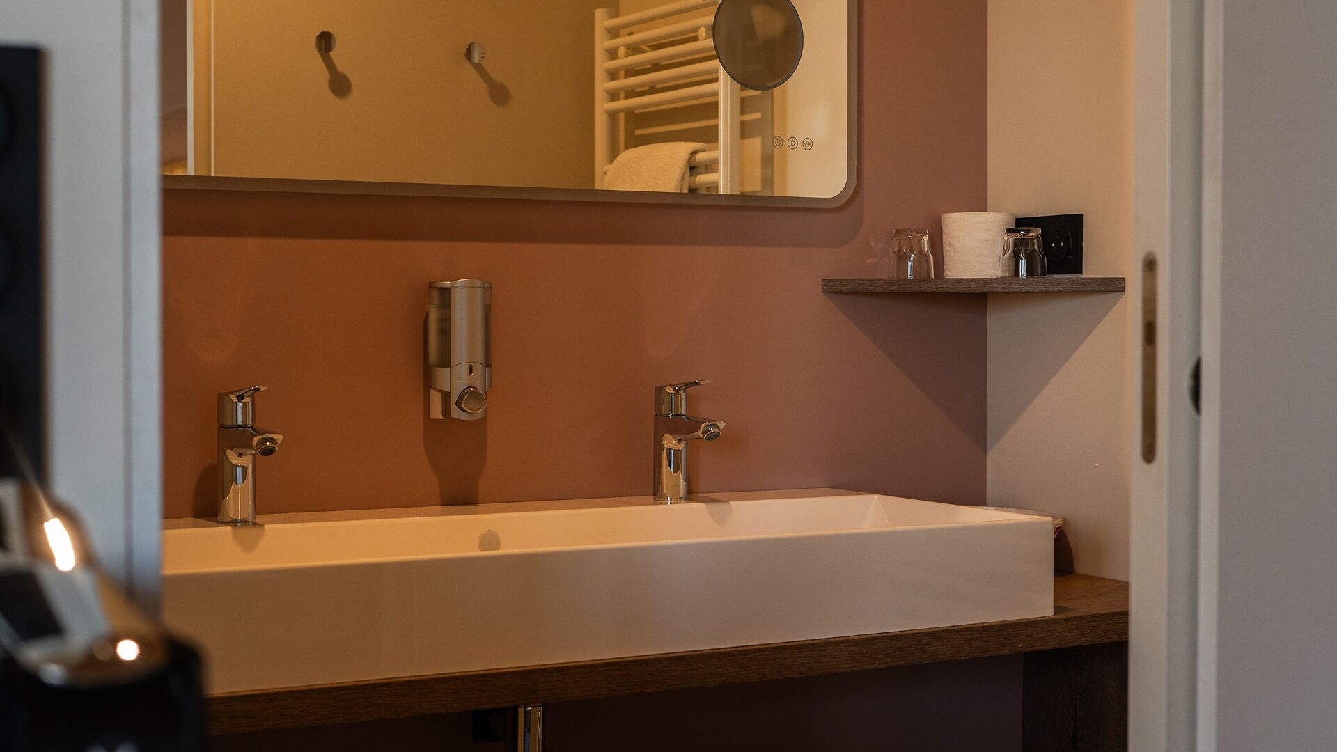 A bathroom with a large sink, two faucets, a mirror, a towel rack, and a shelf with toiletries.