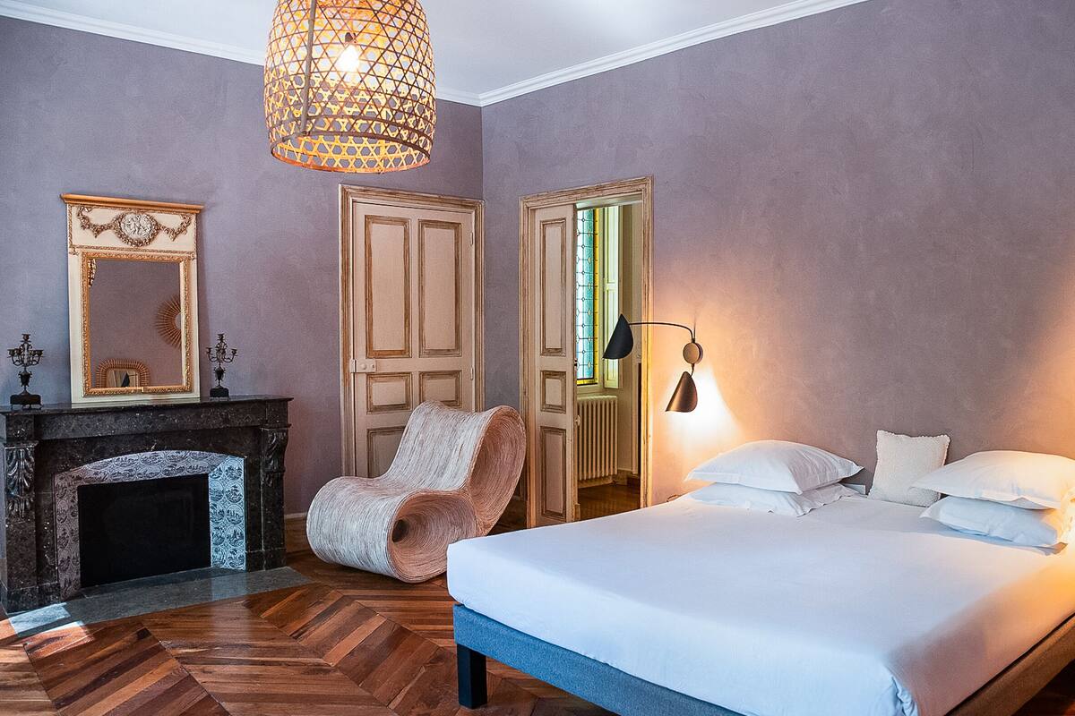 A bedroom with a bed, a chair, a fireplace, and a mirror.