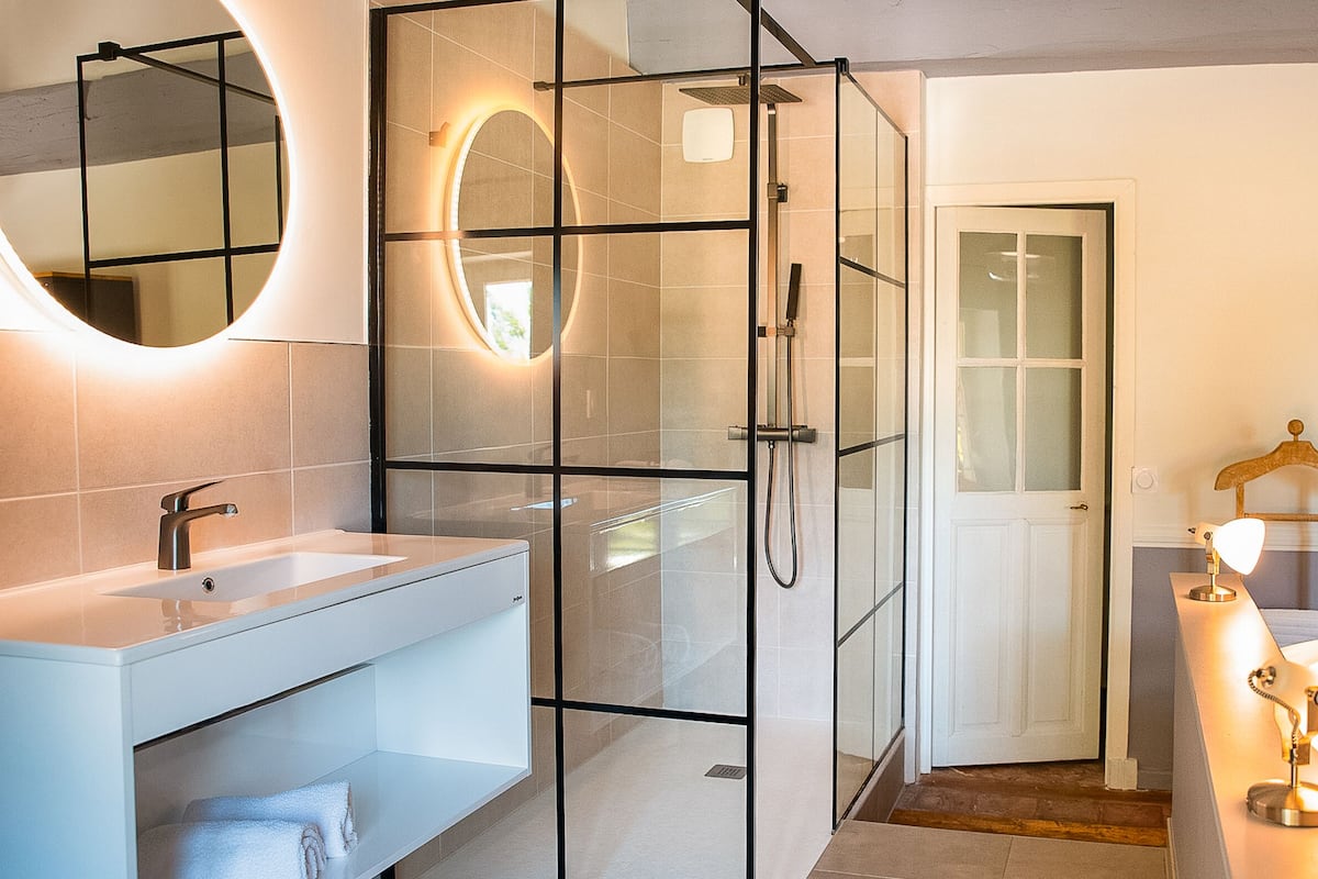 A modern bathroom with a glass-enclosed shower, a vanity with a mirror, and a bedside table with a lamp.