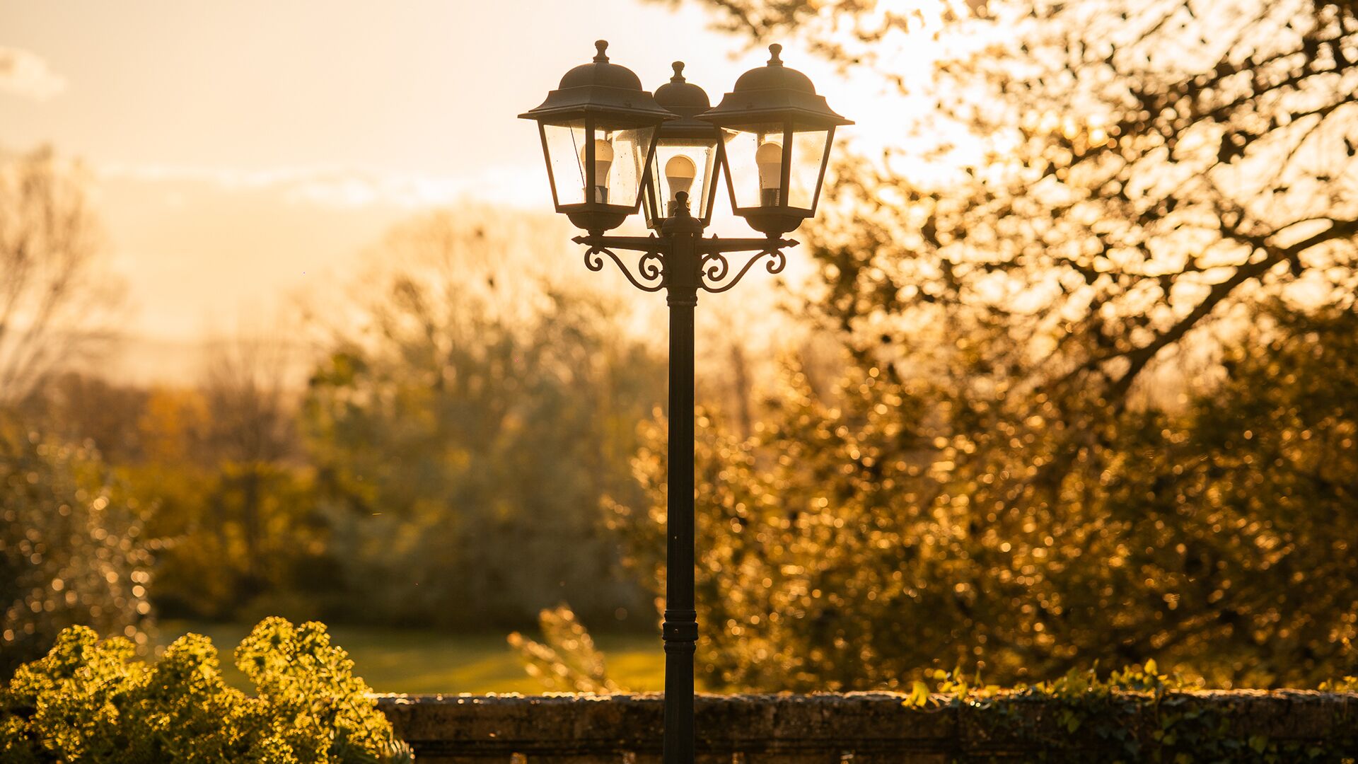 Um candeeiro de rua com três luzes, com um fundo desfocado de árvores e um muro de tijolos.