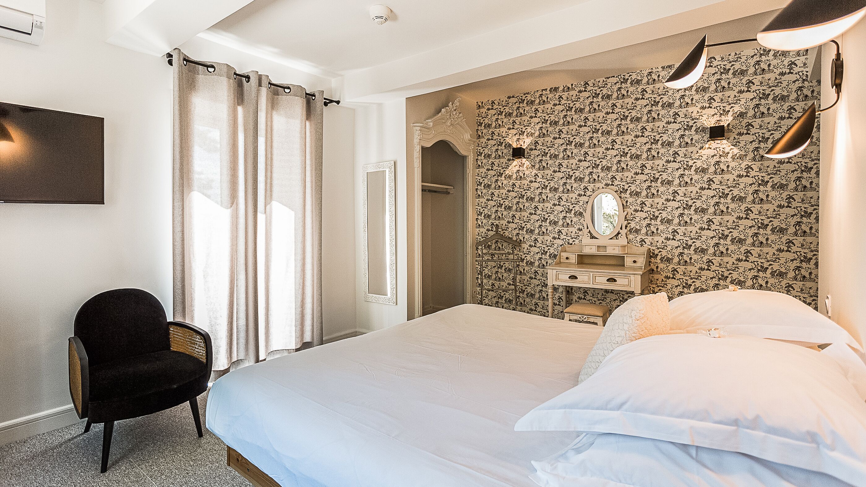 A bedroom with a bed, a chair, a dresser, a mirror, and a wall with a floral pattern.