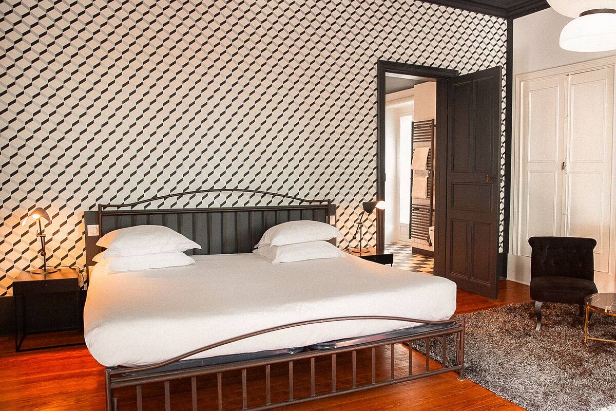 A room with a patterned wall, a bed with white bedding, a small table with cups, and a fireplace.