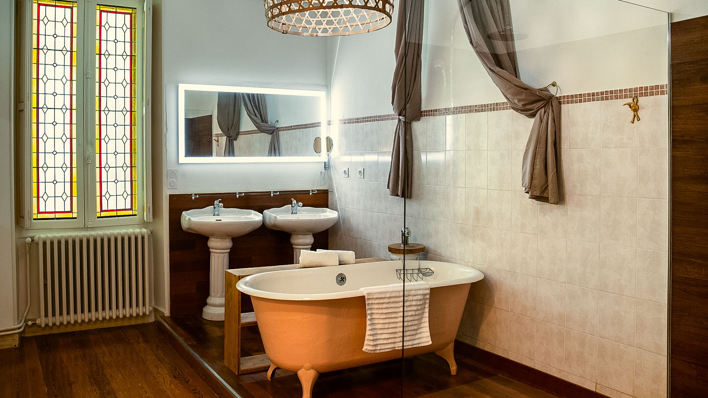 Een badkamer met een pootbad, twee wastafels op voet, een spiegel boven de wastafels, een radiator en een raam met glas-in-lood.