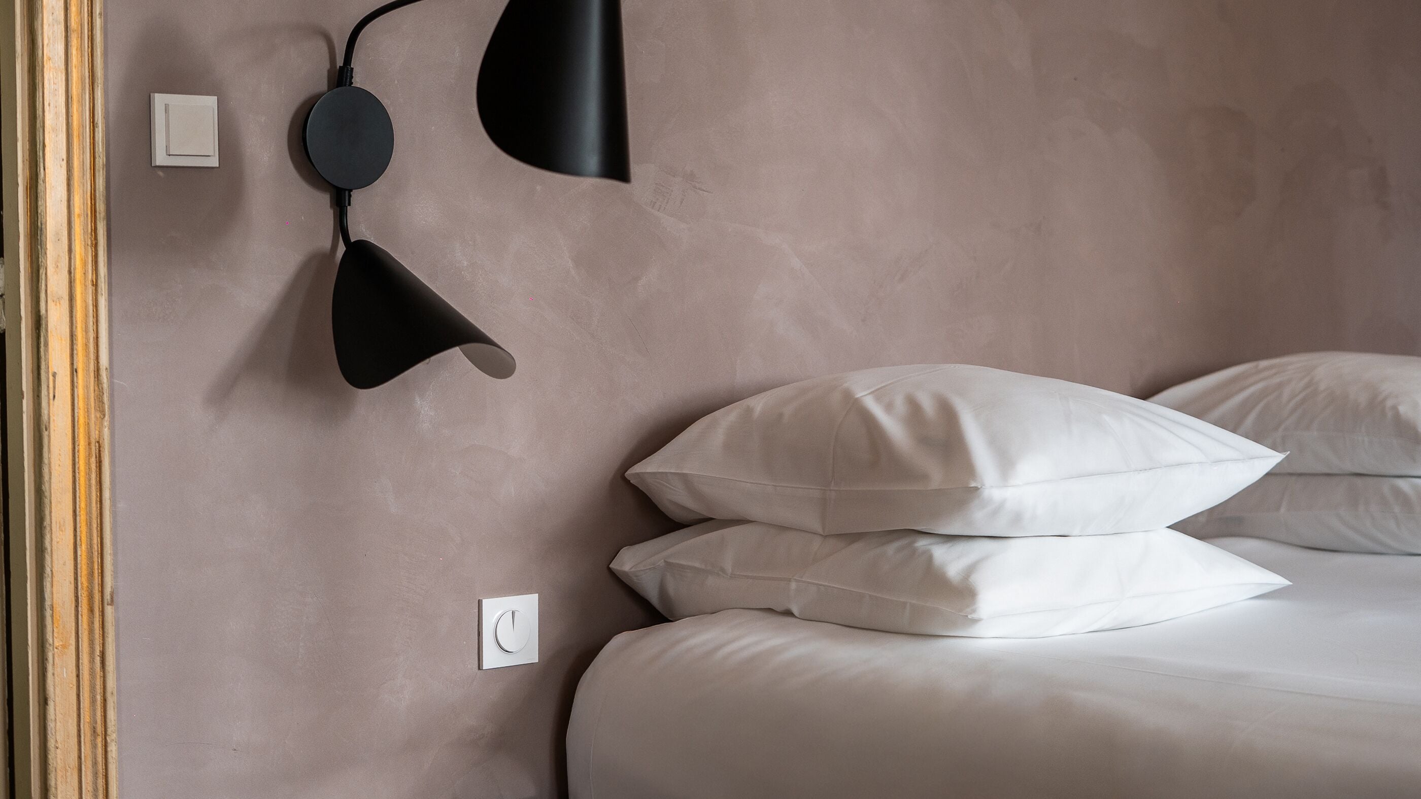 A bedroom with a bed, two pillows, a wall-mounted light fixture, and a power outlet.