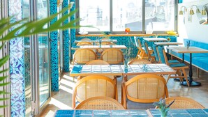 Een restaurant met blauwe behang, rieten meubels en grote ramen met uitzicht op de stad.