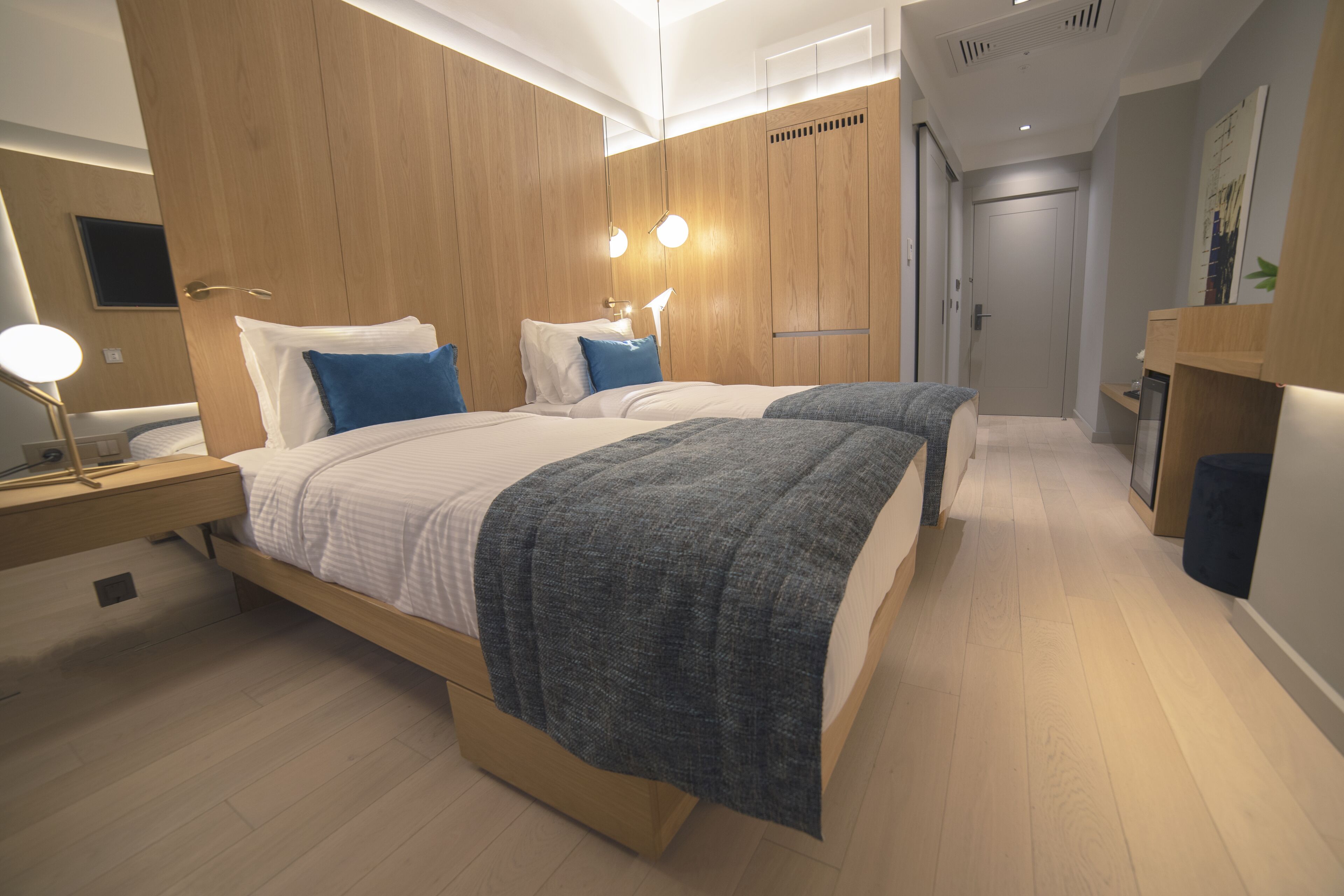 A modern hotel room with two beds, wooden paneling, a desk with a lamp, and a door leading to another room.