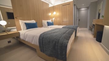 A modern hotel room with two beds, wooden paneling, a desk with a lamp, and a door leading to another room.