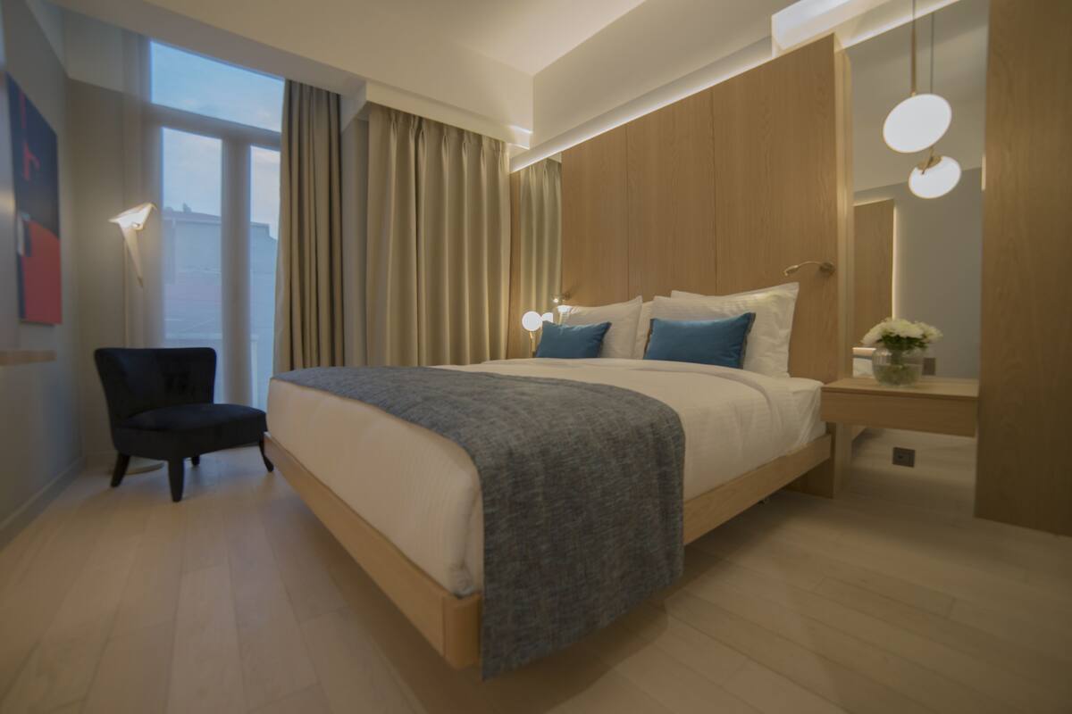 Una habitación de hotel moderna con una cama grande, una silla, una mesita de noche con lámpara y vistas a la ciudad por la ventana.