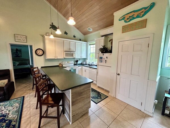 A kitchen with white cabinets, a central island, wooden chairs, a clock, and a sign on the wall.