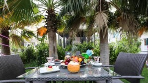 Un área de comedor al aire libre con mesa de vidrio y sillas de mimbre, rodeada de palmeras y follaje.