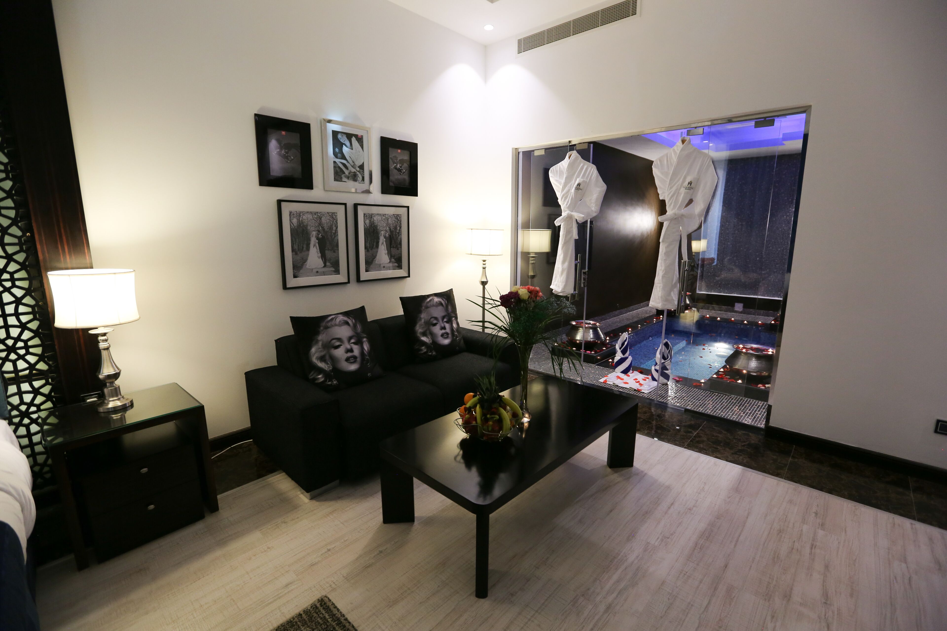 A modern hotel room with a black sofa, a coffee table with fruit, framed pictures on the wall, and a view of a pool through a glass door.