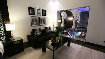 A modern hotel room with a black sofa, a coffee table with fruit, framed pictures on the wall, and a view of a pool through a glass door.