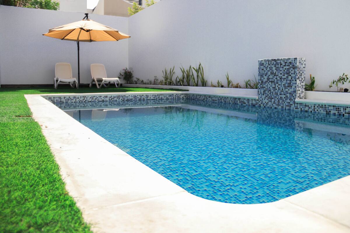 A swimming pool with a tiled edge, two lounge chairs, and a small umbrella.