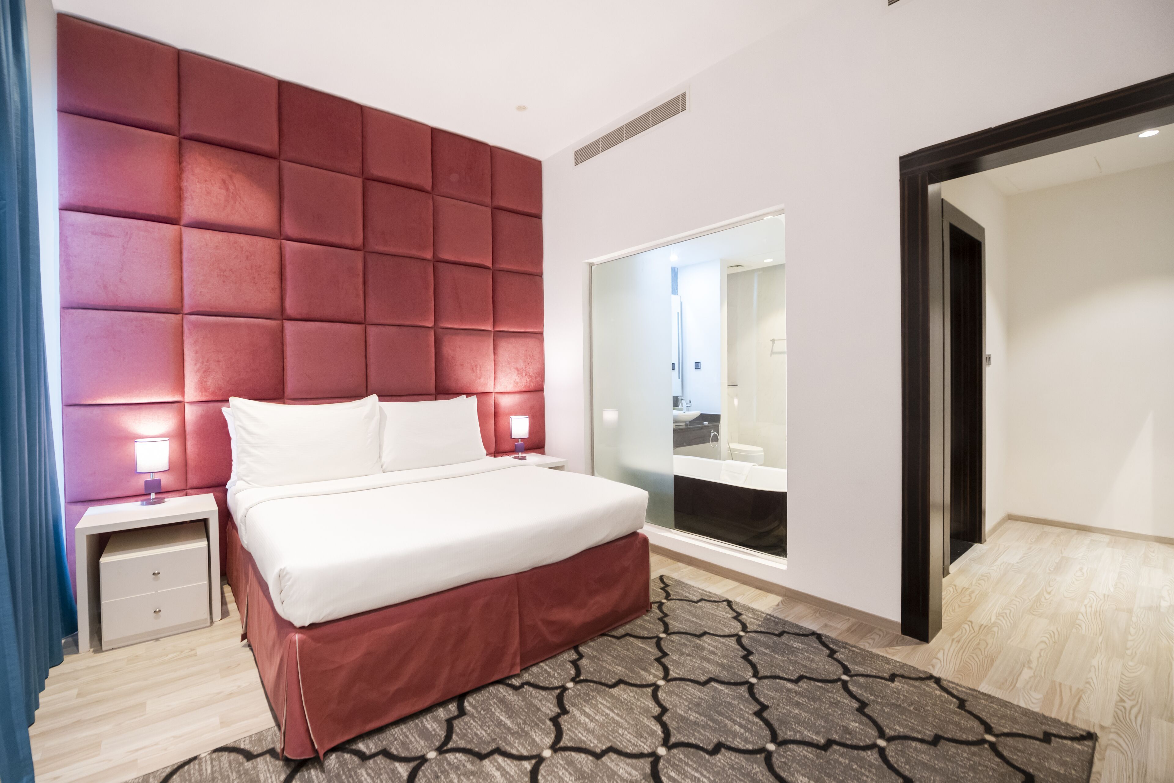 A modern hotel room with a large bed, a red headboard, a bedside table with a lamp, and a large mirror.