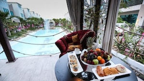 Una mesa de comedor con frutero, pasteles y café, con vista a una piscina y una zona ajardinada con cascada.