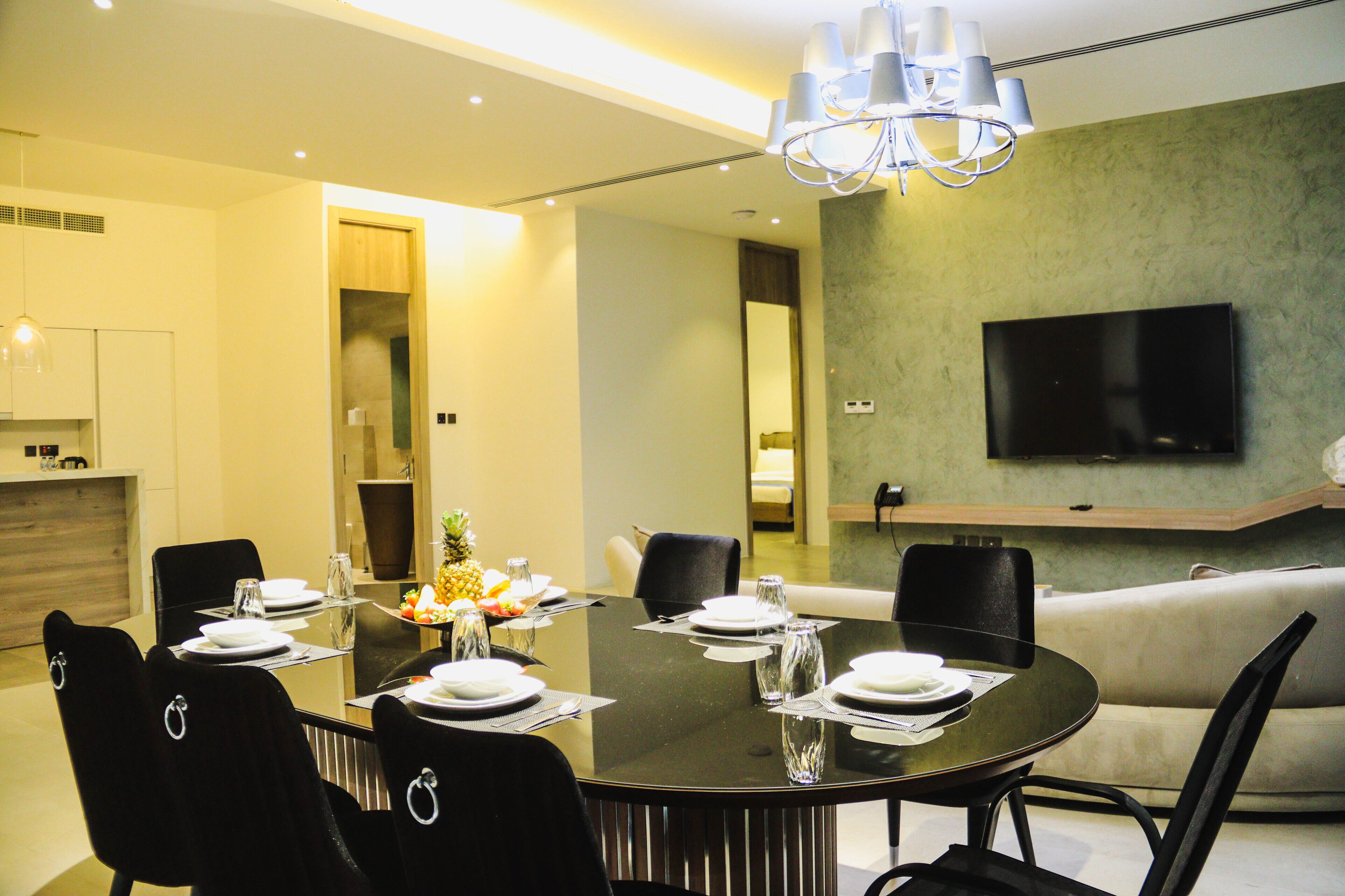 A modern dining area with a round table set for six, surrounded by black chairs. A flat-screen TV is mounted on the wall, and a sofa is visible in the background.