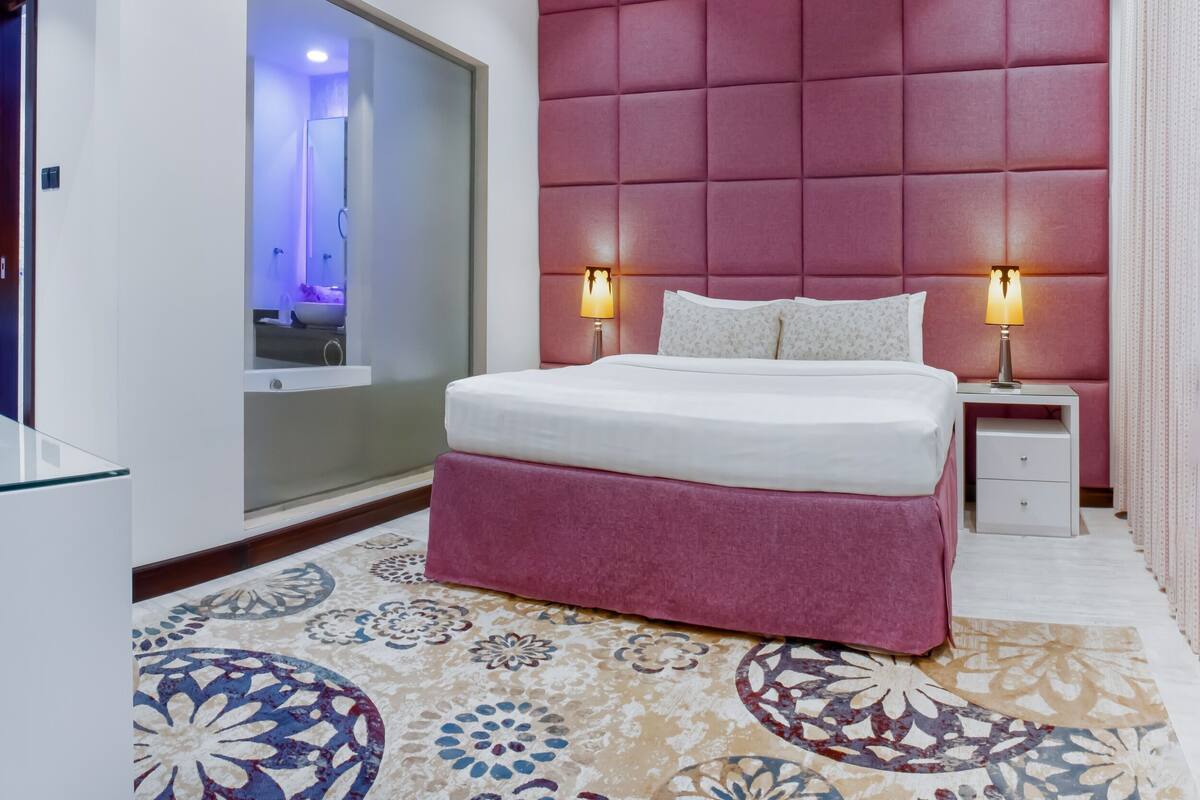 A modern hotel room with a large bed, two bedside lamps, a red padded headboard, and a patterned carpet.