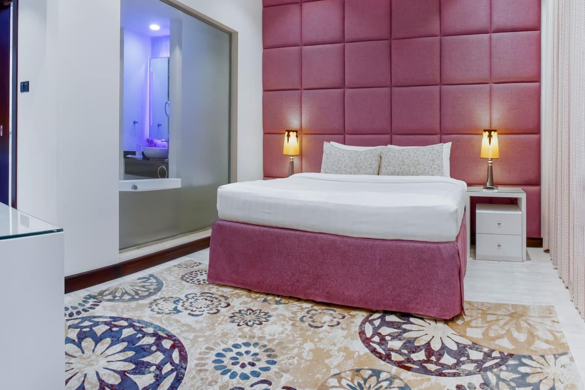 A modern hotel room with a large bed, two bedside lamps, a red padded headboard, and a patterned carpet.