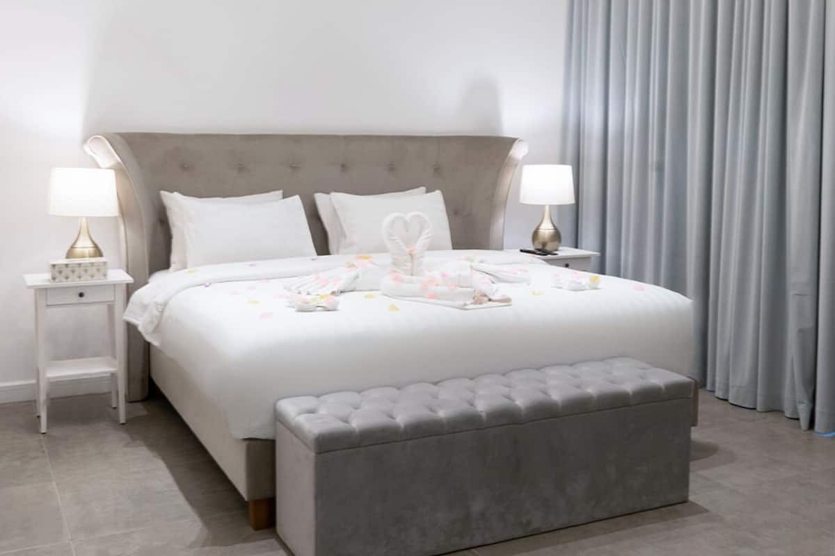 A modern bedroom with a large bed, two bedside tables, a grey upholstered bench, and a grey curtain.