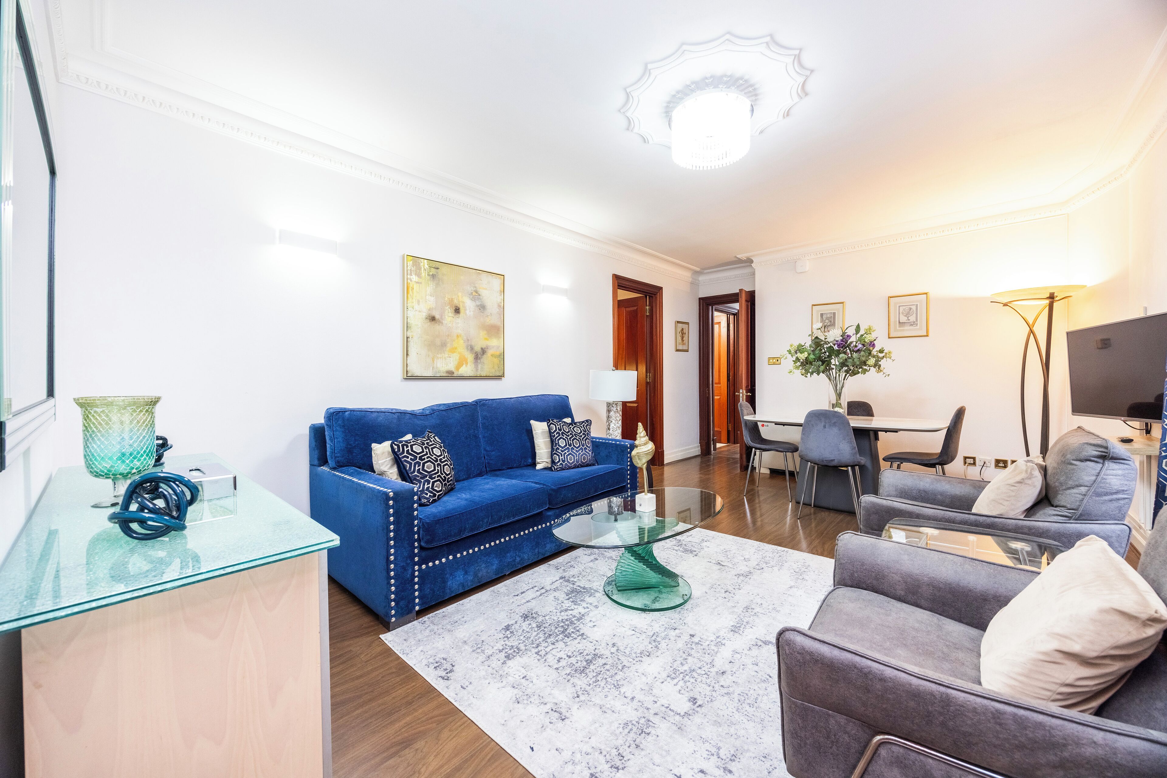 A modern living room with a blue sofa, a grey armchair, a glass coffee table, and a white rug.