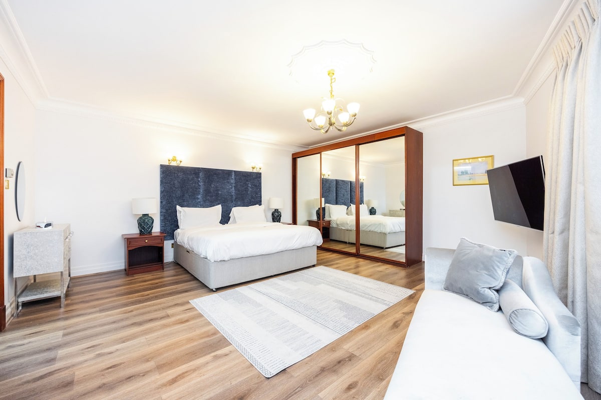 A modern bedroom with a large bed, a sofa, a mirror, a TV, and a chandelier.