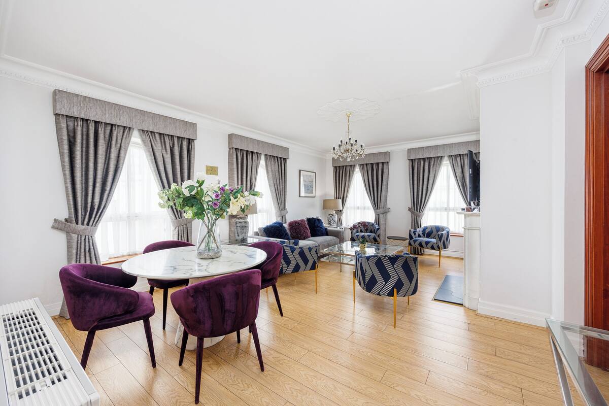 A spacious living room with a round dining table, purple upholstered chairs, a sofa, and patterned armchairs.