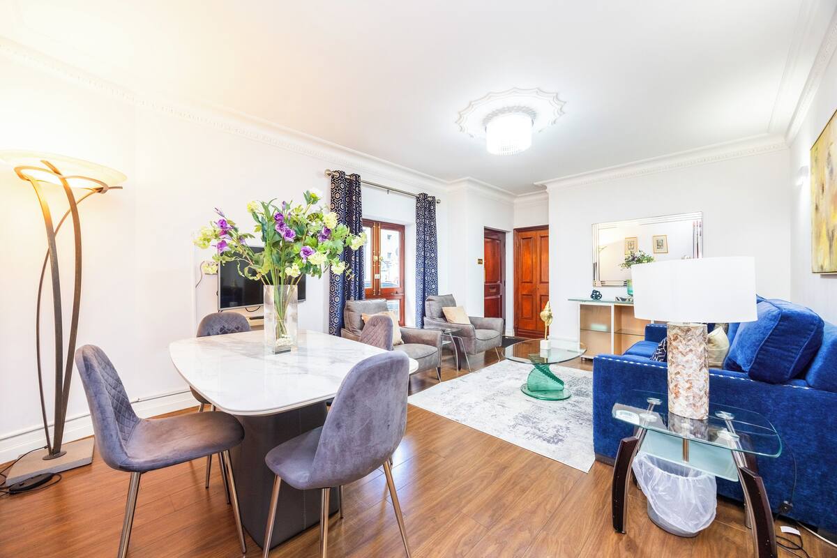 A modern living room with a dining area, featuring a marble table, grey upholstered chairs, a blue sofa, and a decorative vase with flowers.