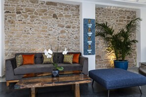 A modern living room with a stone wall, a grey sofa with patterned cushions, a wooden coffee table with orchids, and a blue potted plant. - Suite Azur Hotel (Essaouira)