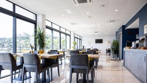 A modern dining area with tables and chairs, large windows, and a view of the city. - Hôtel Le Bastia (Bastia)