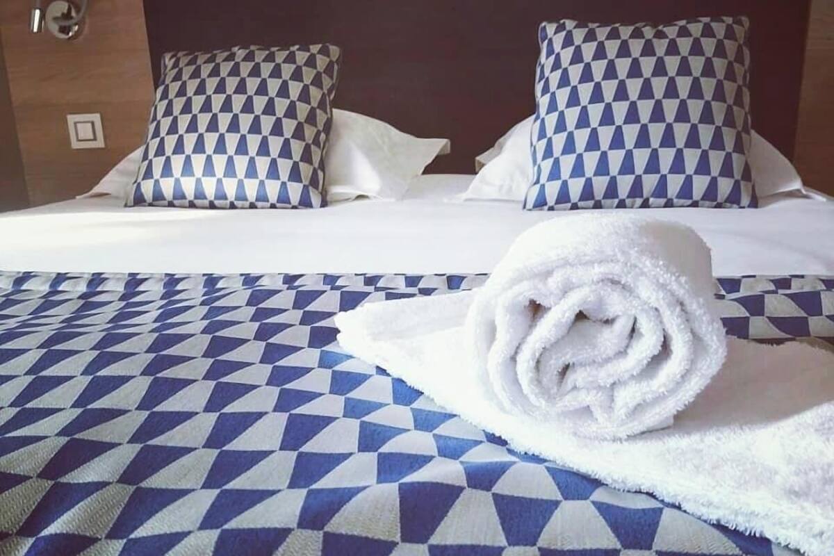 A hotel room with a bed featuring a blue and white checkered bedspread, two matching pillows, and a rolled white towel on the bed.