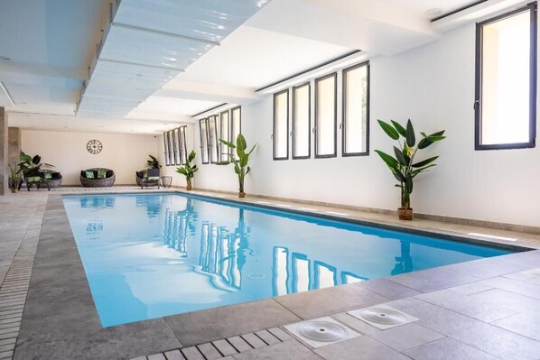 An indoor swimming pool with clear blue water, surrounded by tiled flooring and potted plants. - Hôtel Le Bastia (Bastia)