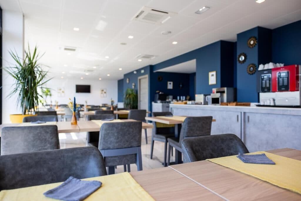 Een moderne eetruimte met tafels en stoelen, een bar met een koffieapparaat en een grote potplant.