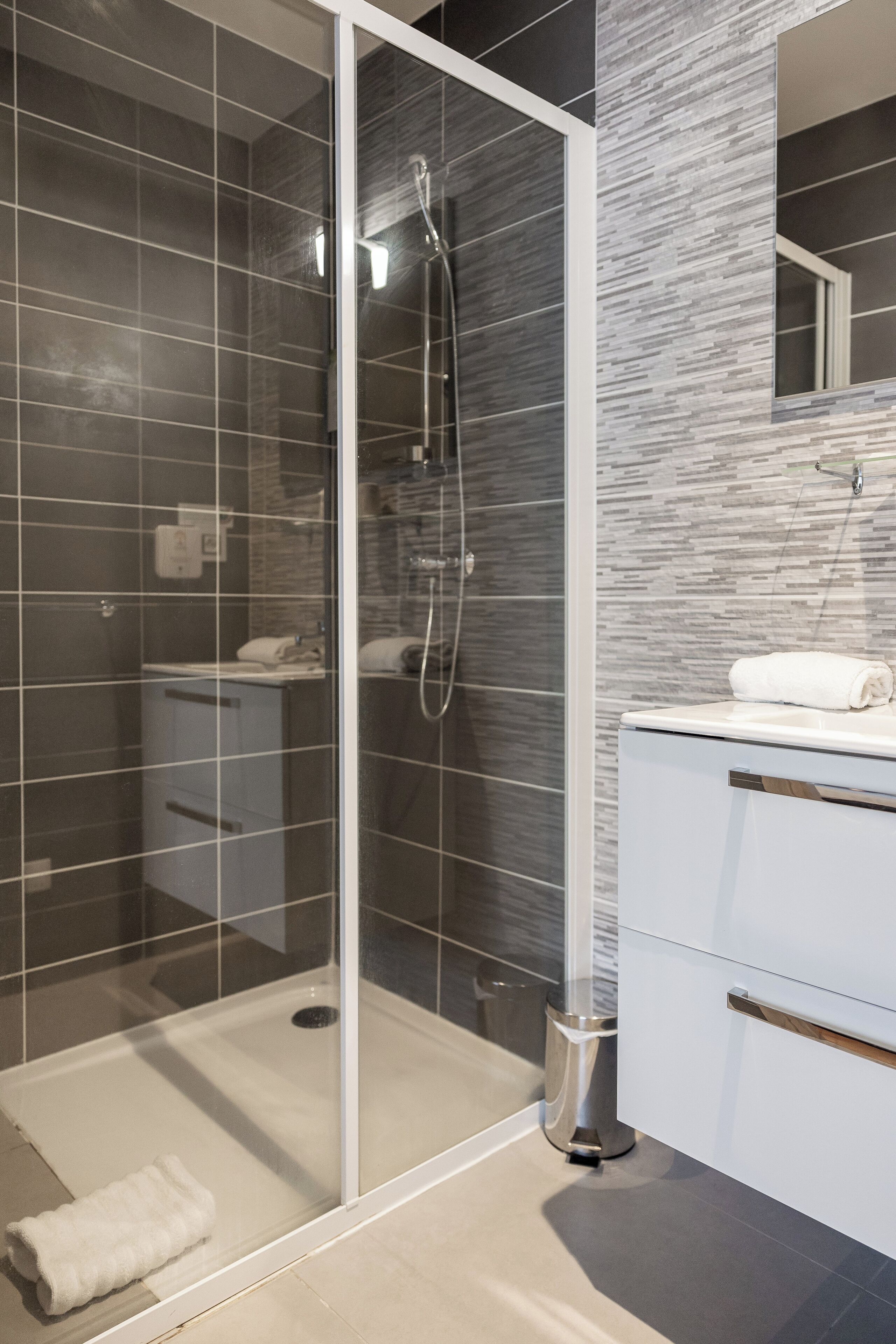 Een moderne badkamer met een douche met glazen wand, een witte wastafel met spiegel en een betegelde vloer.