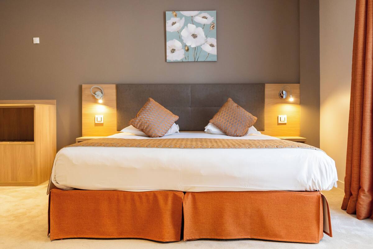A neatly made bed with a white and orange bedspread, two matching pillows, and a wooden headboard. There are two bedside lamps and a wall-mounted artwork above the bed.