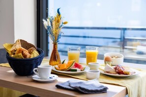 A breakfast setup with bread, fruits, eggs, and juice on a table by a window with a view. - Hôtel Le Bastia (Bastia)